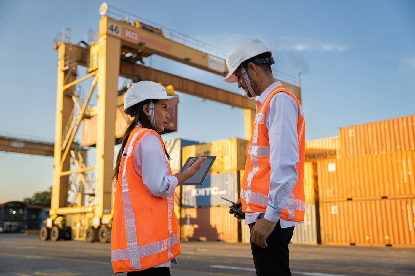 Two workers in hard hats and high-visibility vests reviewing plans beside shipping containers